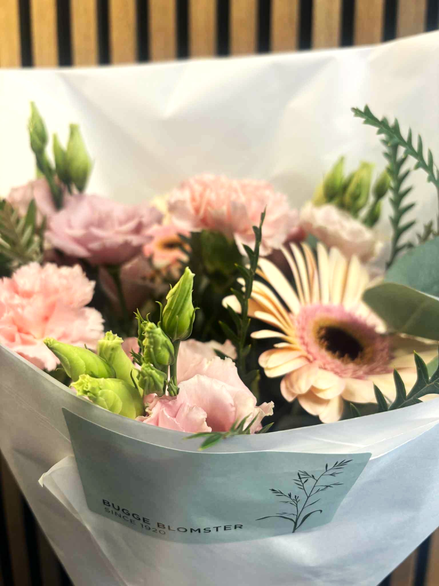 Buketten er bundet med blomster i sarte pastelfarver og pakket ind i hvidt papir. 
