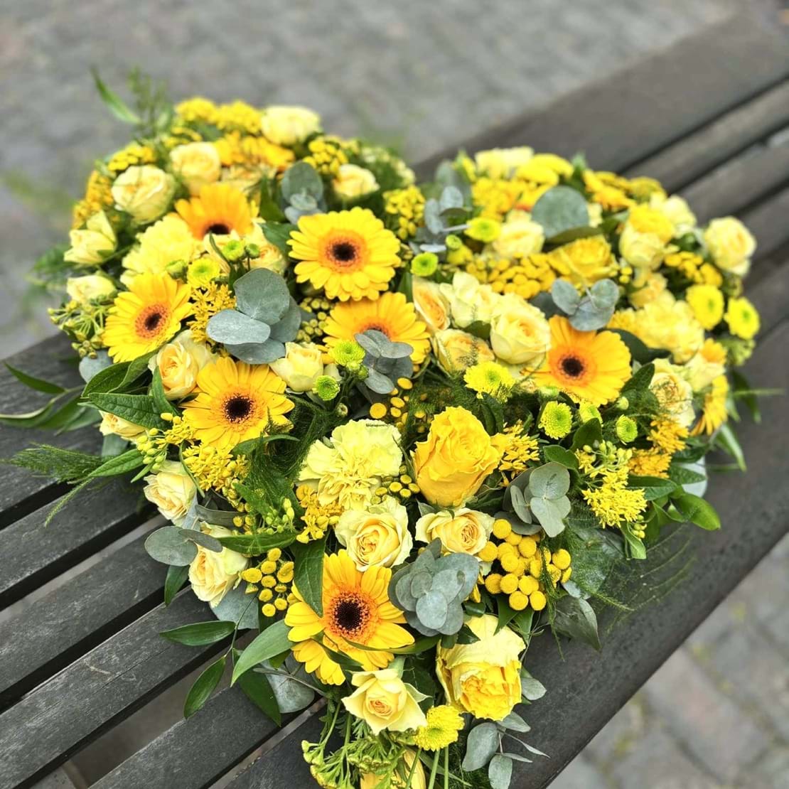 Begravelseshjerte med lyse og skarpe gule blomster i forskellige størrelser. Den ligger på en sort træbænk.