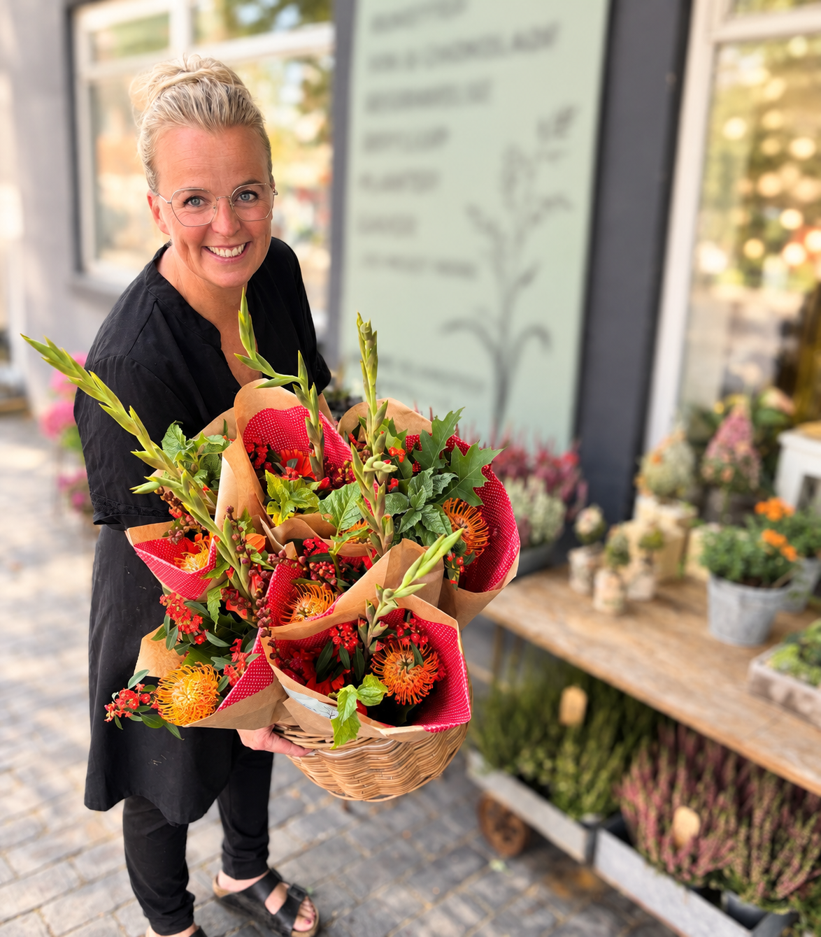 Florist fra Bugge Blomster, står foran butikken og holder en kurv med flere store, smukke indpakkede buketter af friske blomster.