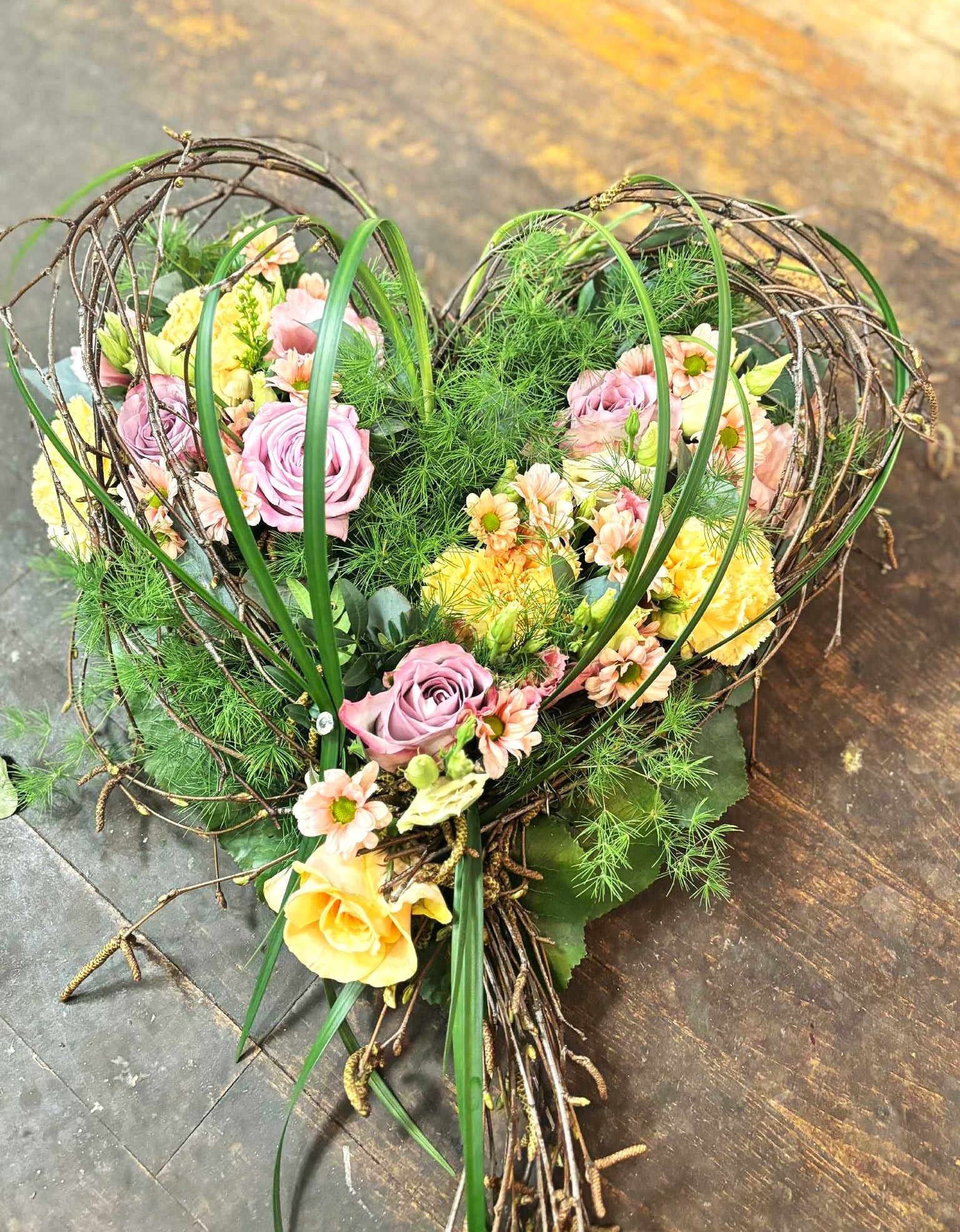 Hjerte bundet med grønne naturmaterialer og stukket med blomster i lyse pastel nuancer. 