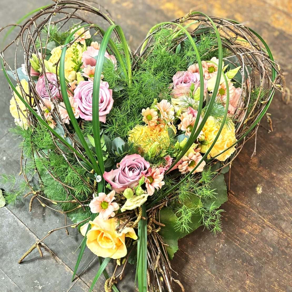 Hjerte bundet med grønne naturmaterialer og stukket med blomster i lyse pastel nuancer.