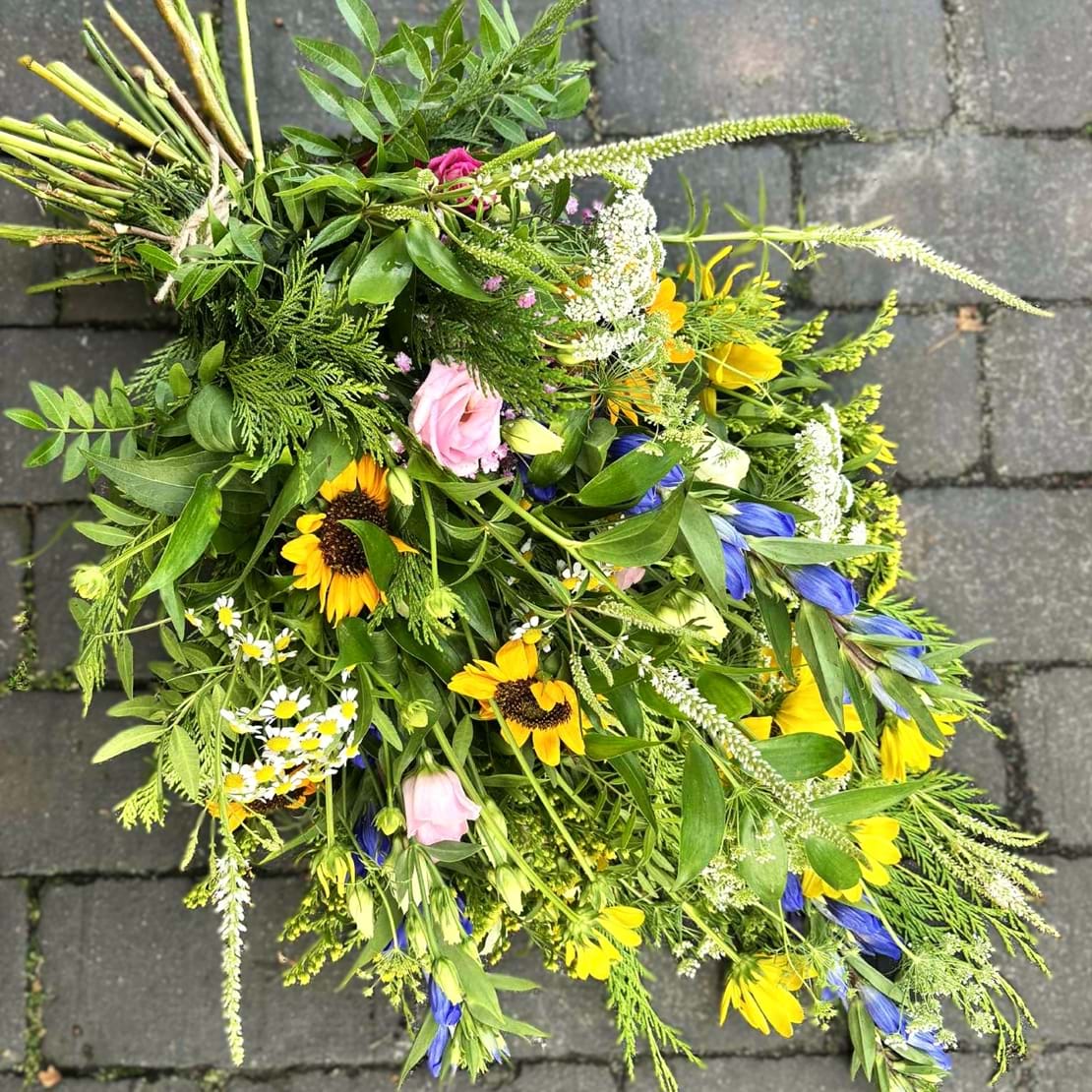 Bårebuket med blomster i friske farver som gul, blå og lysegrøn. Den er bundet i naturligt og livligt udtryk.