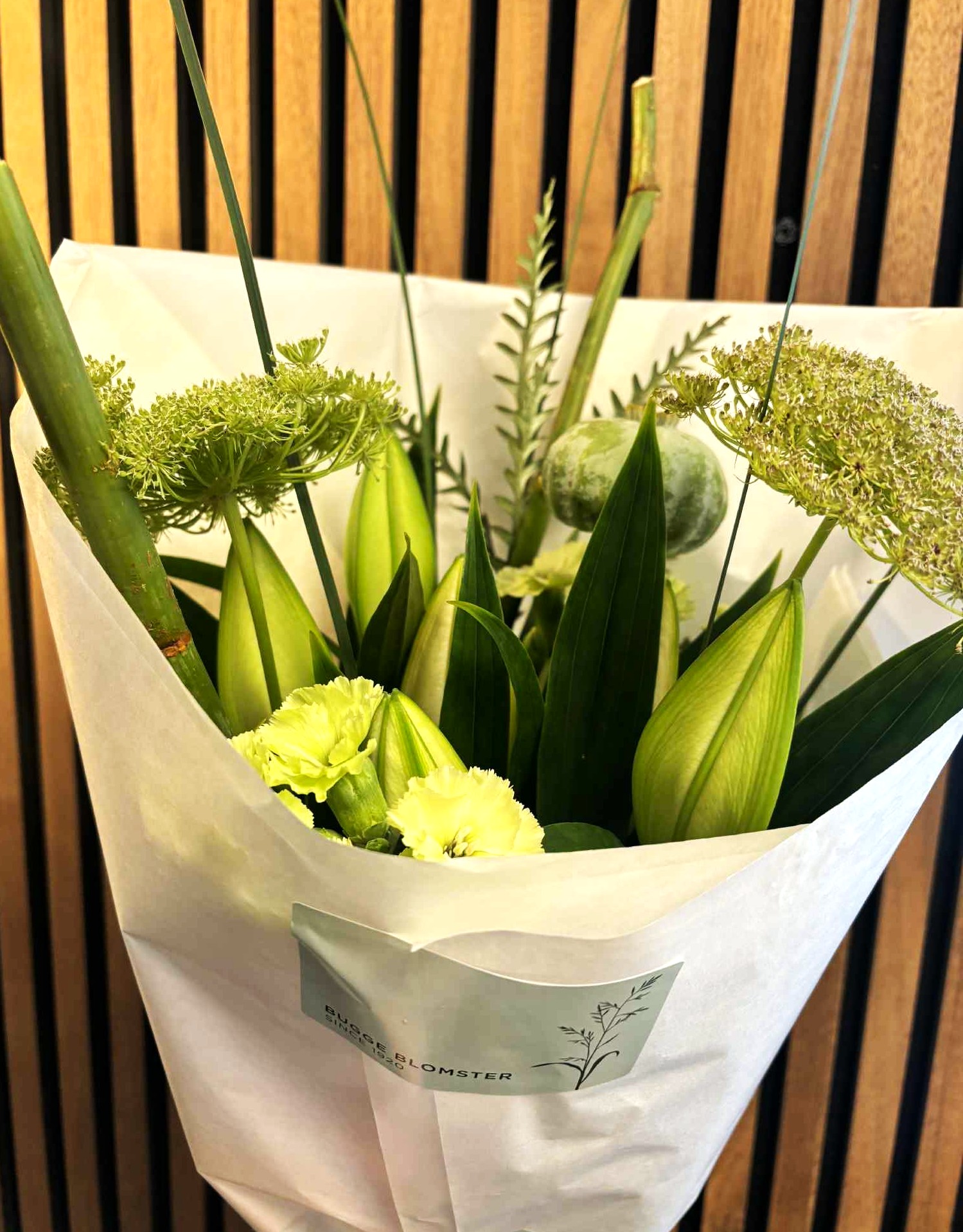 Buket bundet med grønne blomster. Buketten er pakket ind i hvidt papir og påsat et klistermærke fra Bugge Blomster.
